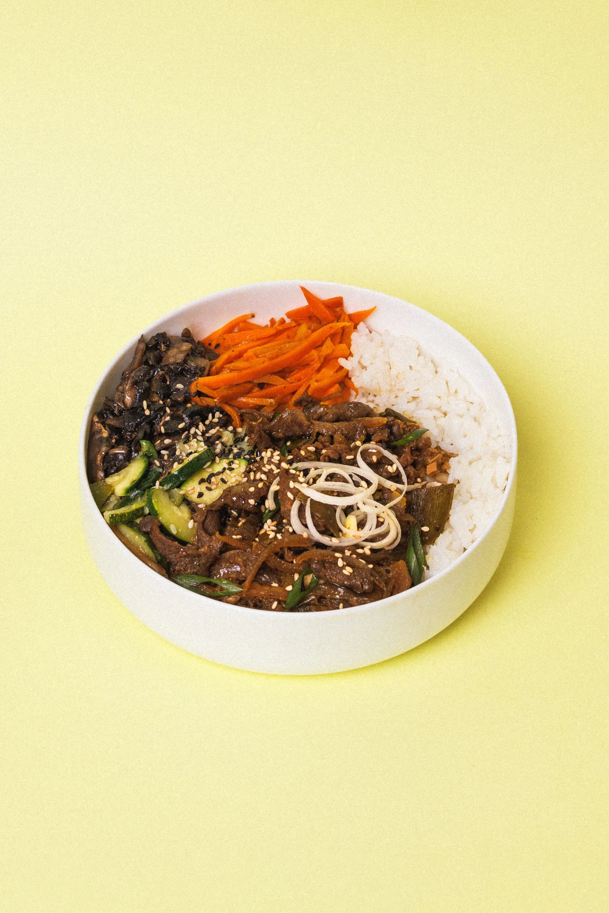 A white round bowl filled with  Korean bulgogi  rice dish on a solid pale yellow background. The dish includes white rice, stir-fried vegetables, dark mushrooms, and garnishes of shredded carrots, sesame seeds, and sliced scallions.