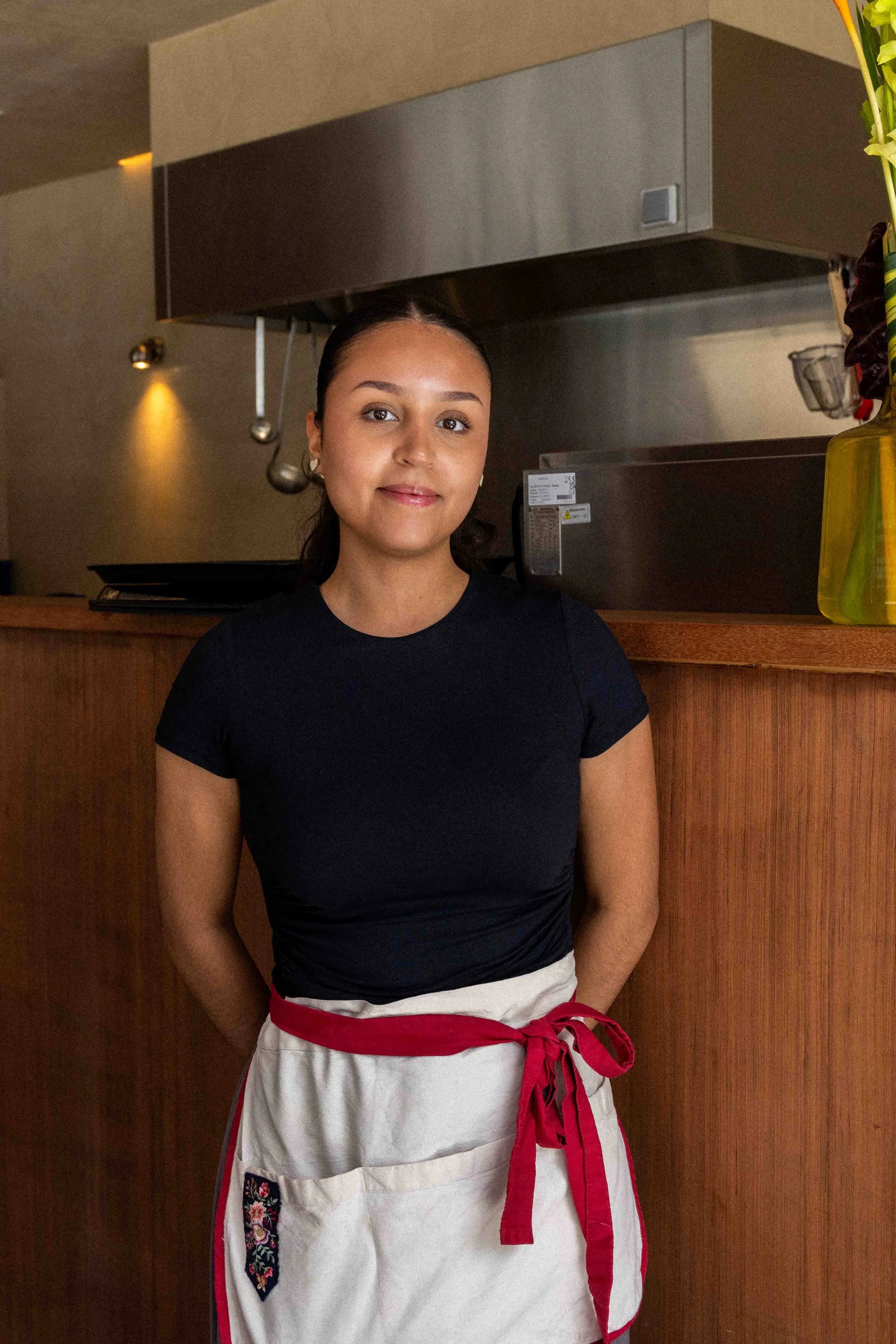 A woman wearing a black short-sleeve t-shirt and a white apron with a red waist tie, standing in a kitchen environment.